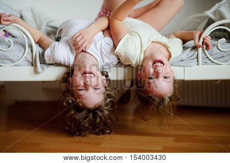 The children boy and girl naughty on the bed in the bedroom. They lie side by side their heads hanging off the bed. Children is very amused.