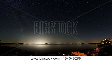 Panorama of the night sky above lake Mjörn in Sweden. The Milky Way is visible on the left side and the bright light of large cities on the opposite side of the lake. In the foreground is the rocky coastline.