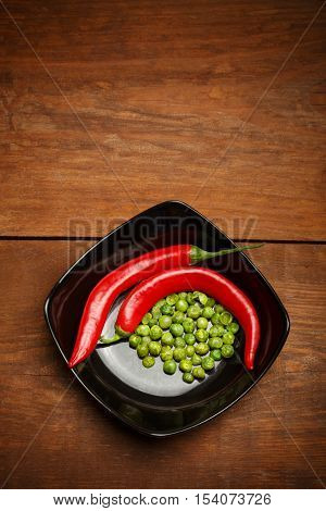 Red hot chilli peppers and crispy roasted green peas snacks coated in wasabi lying in ceramic black plate on brown wooden table. Copyspace. Top view.