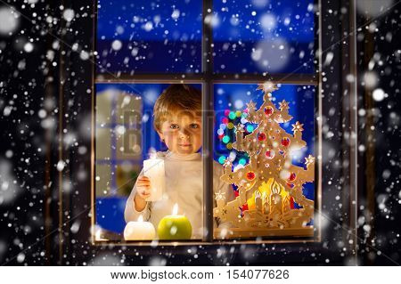 Beautiful little kid boy, standing by window at Christmas time and holding candle. Child waiting for Santa Claus. and gifts. With colorful lights from Christmas tree on background.