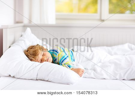 Carefree little kid boy sleeping in his white bed in colorful nightwear near big window with green and yellow autumn foliage. Funny happy child playing and smiling. Family, vacation, childhood concept