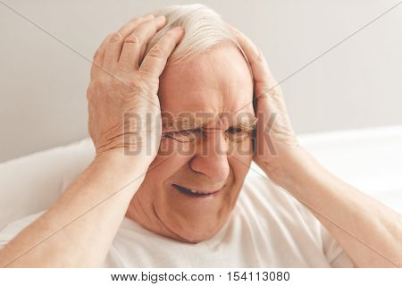 Handsome old man is having a headache. He is holding his hands on head lying in bed in hospital