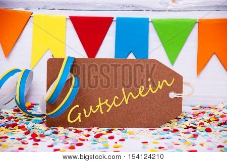 Brown Label With German Text Gutschein Means Voucher. Party Decoration Like Streamer And Confetti. White Wooden Background. Greeting Card For Celebrations