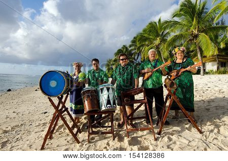 Group portrait of Polynesian Pacific Islanders band plays Tahitian music on tropical beach with palm trees in the background.