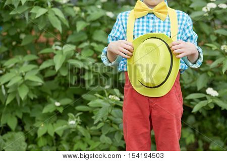 Body of little boy dressed in dance suit holding yellow hat pressed to belly standing outdoor.