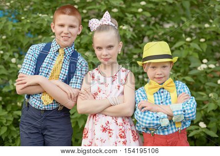 Two boys in checkered shirts and trousers and girl in flowery gown stand with arms folded on chests.