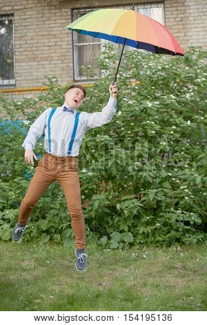 Smiling teenage boy jumps holding rainbow coloured umbrella at lawn near house.