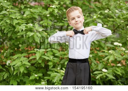 Little boy dressed in dance suit adjusts bow standing outdoor.