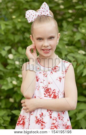 Girl in flowery gown touches earring standing outdoor against green bushes.