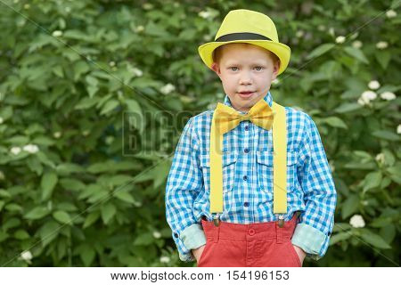 Boy in yellow hat with hands in pockets against green bushes.