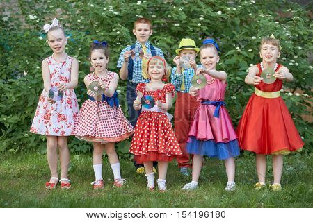 Five smiling little girls and two boys dressed in dancing suits pose at grassy lawn with cd in hands.