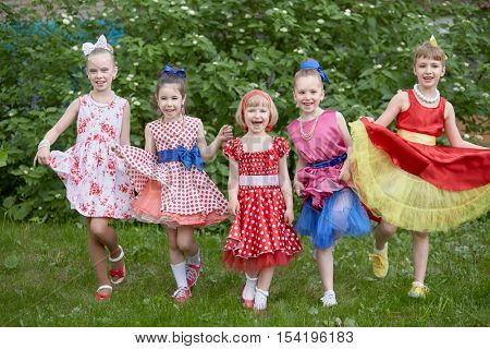 Five smiling little girls dressed in dancing suits pose at grassy lawn.