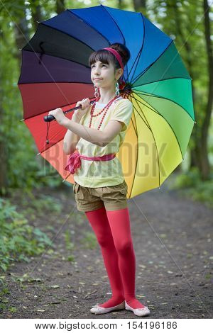 Girl with rainbow coloured umbrella stands in summer park looking upwards.