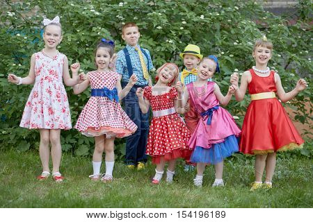 Five smiling little girls dressed in dancing suits and two boys pose at grassy lawn.
