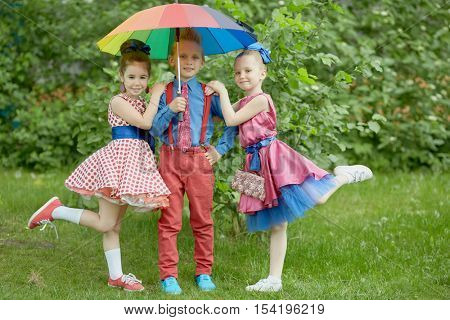 Boy in red trousers and blue shirt stands with two girls under coloured rainbow umbrella at grassy lawn.