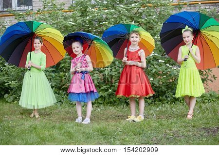Four girls in dance suits stand under coloured rainbow umbrellas outdoor at lawn.
