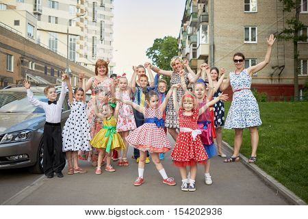 Group of eleven children and four woman stands posing at sidewalk.