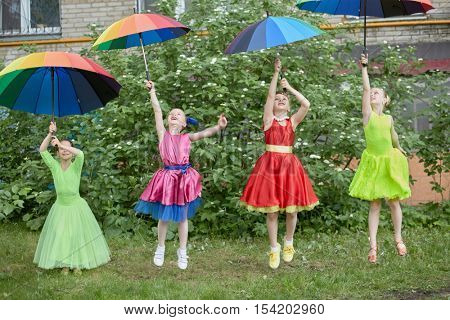 Four girls in dance suits jump throwing up coloured rainbow umbrellas outdoor at lawn.