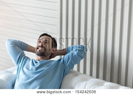 Feeling great. Nice cheerful bearded man holding his hands behind the head and smiling while being in a wonderful mood