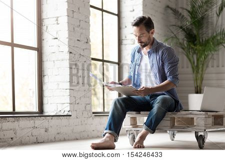 Working at home. Serious good looking adult man holding some documents and reading them while being at home