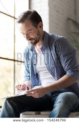 New ideas. Creative delighted handsome man leaning forward and looking at his notes while writing something there