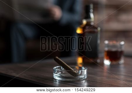 Smoking habits. Selective focus of a cigar lying in the ashtray next to the bottle of whisky with a hard working confident successful businessman sitting in the background