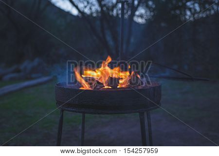 Burning camp fire at dusk in camping site preparing for barbeque or braai outdoors activity in South Africa. Selective focus on fire and firewood very shallow depth of field cold toned image.
