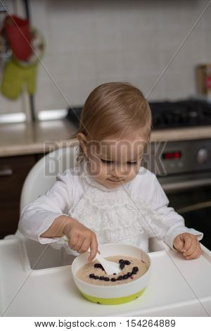 Cute little girl eating healthy porridge with bilberry with plastic spoon while sitting in the high chair at table in the kitchen. Image with selective focus.
