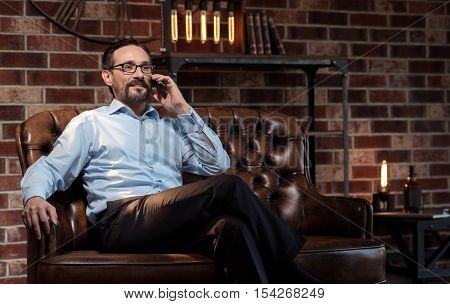 Distance communication. Handsome delighted good looking businessman putting the smartphone to his ear and listening to his interlocutor while being in his study room