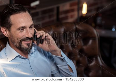 Pleasant conversation. Positive cheerful bearded man listening to his interlocutor and laughing while having a pleasant conversation on the phone