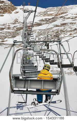 Young snowboarder in a yellow jacket elevating to the mountain top on a chairlift