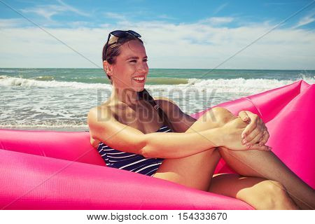 A woman sitting on the pink airbed and placing her hands on the knee, looking squinty at something and beautifully smiling. Marvelous sunny day on the seaside, approaching a wave behind the woman