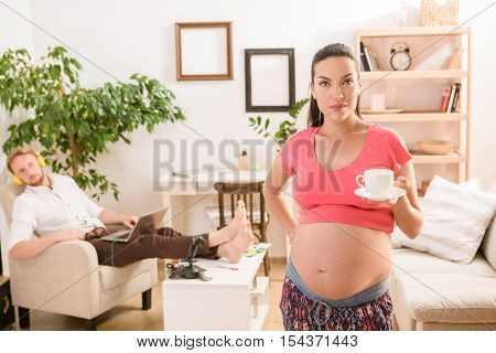 Healthy lifestyle. Picture of beautiful serious pregnant woman offering cup of tea or coffee while staying at home with her husband.