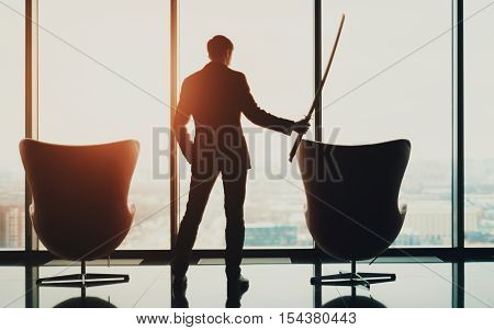 Young serious businessman in office in business suit standing between of two armchairs in front of window and holding katana sword like japanese samurai warrior blurred city top view in background