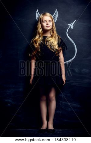 Funny little imp girl. Cute child girl posing with imp horns and tail drawn on a blackboard.