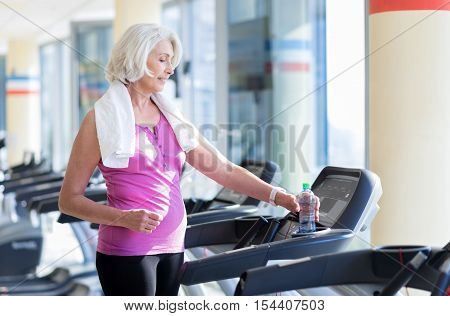Better than yesterday. Pretty positive senior woman smiling and taking bottle of water while having a rest after running on treadmill.