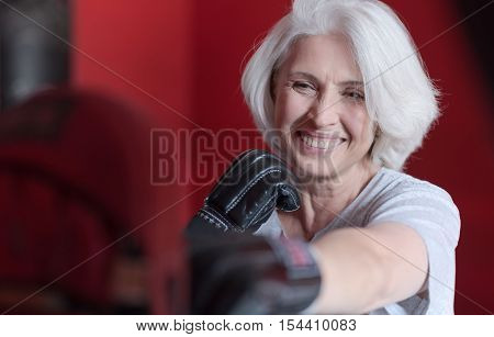 Unstressed moments. Delighted senior charming woman smiling and boxing punching bag while training in a gym.