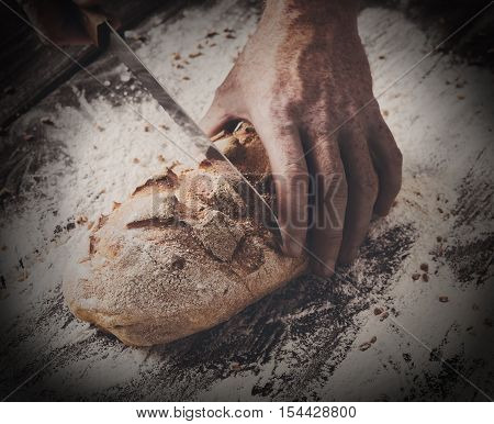 Baking and cooking concept background. Hands of baker closeup cutting bread loaf with knife on rustic wooden table sprinkled with flour. Stained dirty hands of baker, filtered toned image