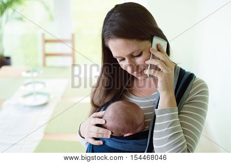 Beautiful young mother with her newborn baby son in sling at home, holding smart phone, making phone call