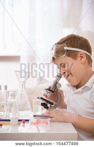 Cute elementary schoolboy looking into microscope at his desk at home. Young scientist making experiments in his home laboratory. Indoors. Child and science. Education concept.