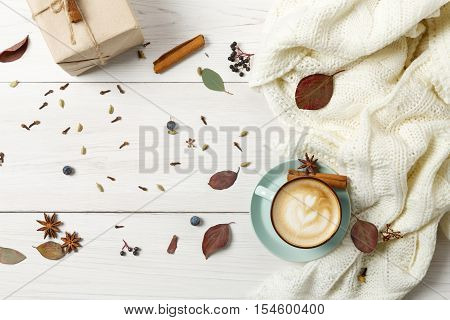 Autumn cappuccino composition. Blue coffee cup top view with foam, cloves, cinnamon, present box and warm sweater at white wood background. Fall hot drinks concept