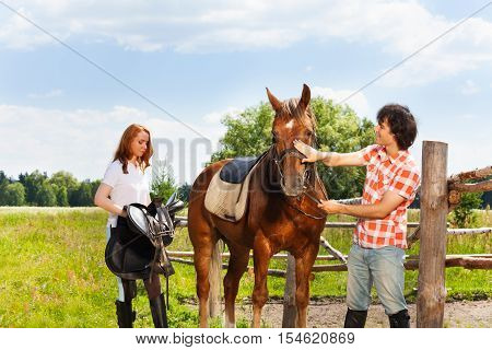 Two happy equestrians, young woman and man preparing their horse for riding at contryside
