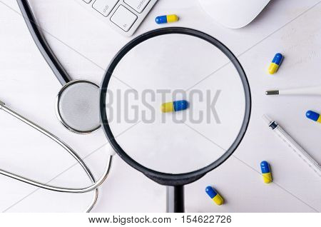 Close-up of magnifying glass and medicine with stethoscope keyboard and mouse. Top view of white wooden working table.