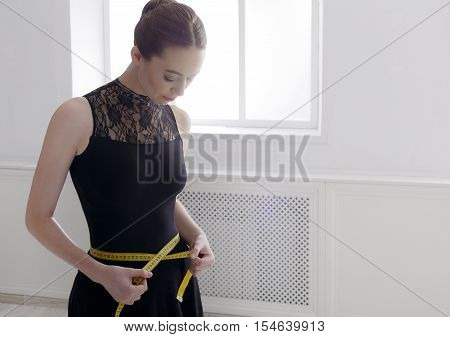 Ballet dancer looks at her perfect waist after diet. Beautiful graceful ballerine in black with measuring tape in light hall. Slimming and weight loss concept, copy space