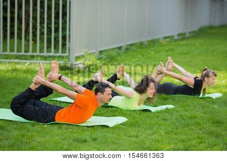 Man and two smiling women practice yoga on grass near fence at summer day