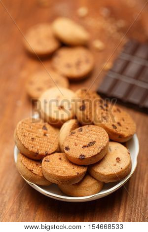 Beautiful tasty biscuit cookies with chocolate chips standing in plate on wooden table with crumbs and chocolate bar at the background. Shallow dof. Focus on foreground.