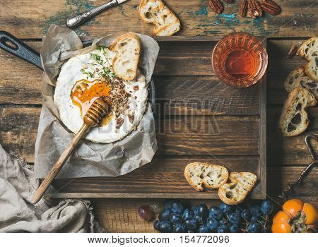Homemade camembert cheese with honey and herbs in small pan, black grapes, persimmon, baguette slices, pecan nuts and glass of rose wine over rustic wooden background, top view, copy space