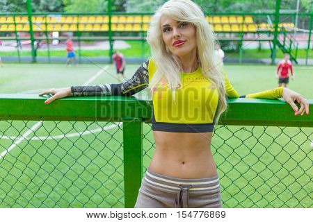 MOSCOW - JUL 16, 2015: Beautiful blonde woman with long hair in top leaned on fence on soccer field