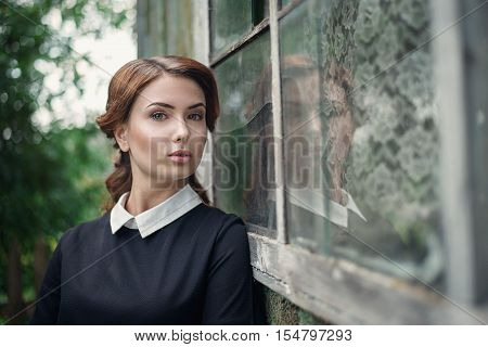 Pensive beautiful young girl in retro style dress standing near the window of old wooden house. Closeup portrait. She is a teacher. Nude makeup.