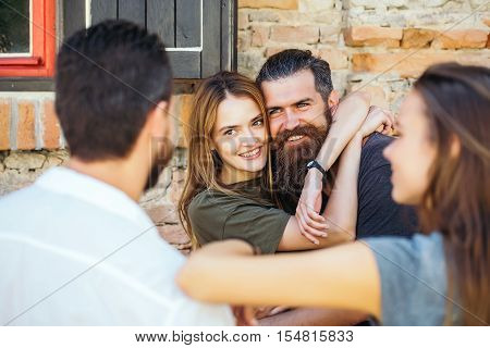 Couple of handsome bearded man with long beard and stylish hair and blonde pretty cute sexy woman or girl embracing and smiling to they friends on summer sunny day on stony wall background